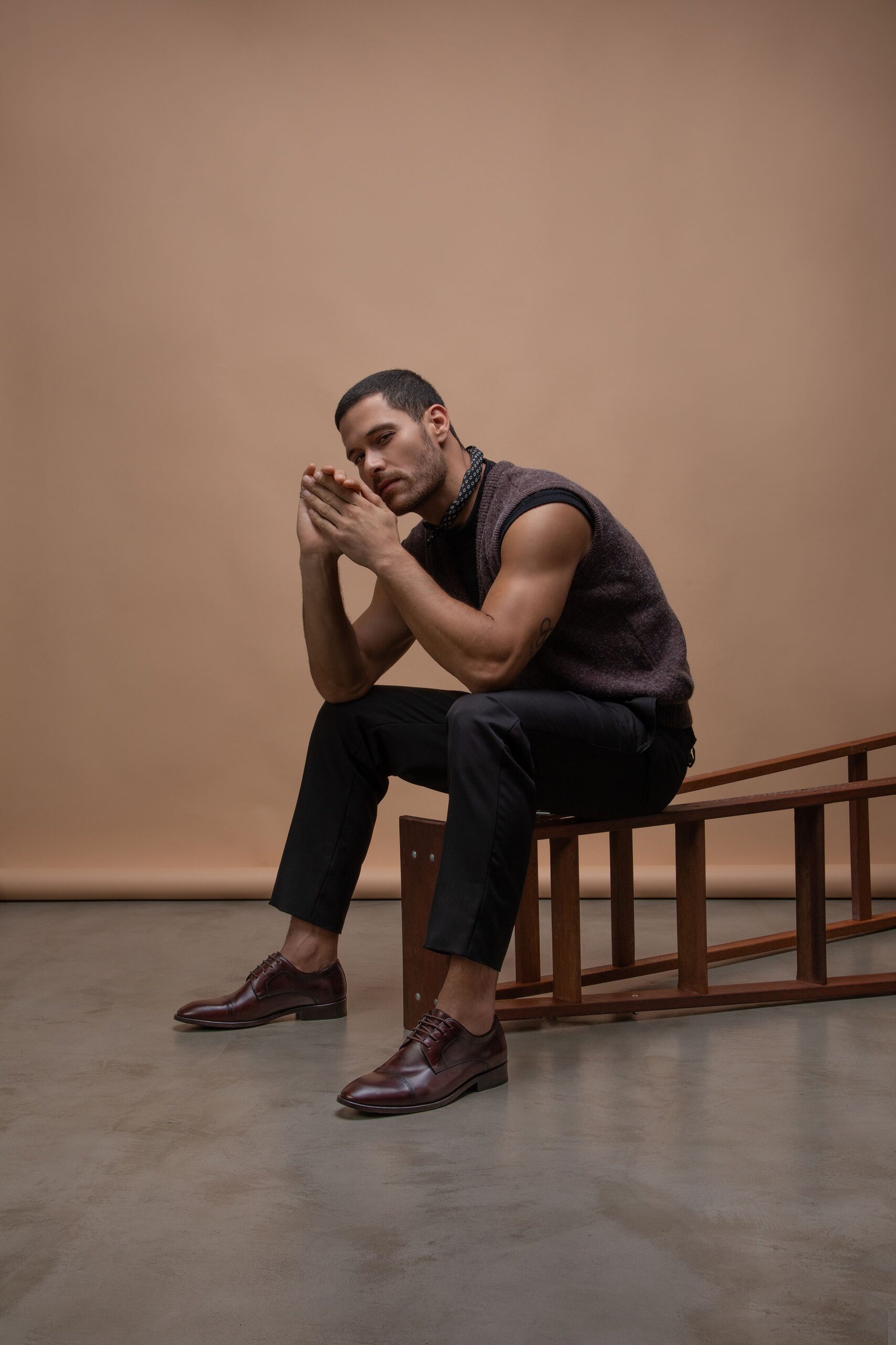 homem com lenço preto no pescoço, usando uma regata de linho marrom, calça social preta, usando um derby douss marrom, sentado em uma escada de madeira deitada no chão
